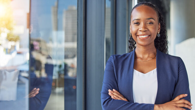 Confident young Black woman in business attire standing with arms crossed outside a modern office building.