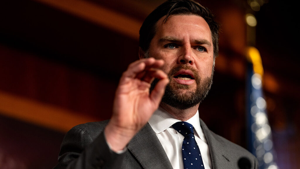 A bearded man in a suit and tie gesturing with his hand while speaking at a podium with a partial view of an american flag in the background