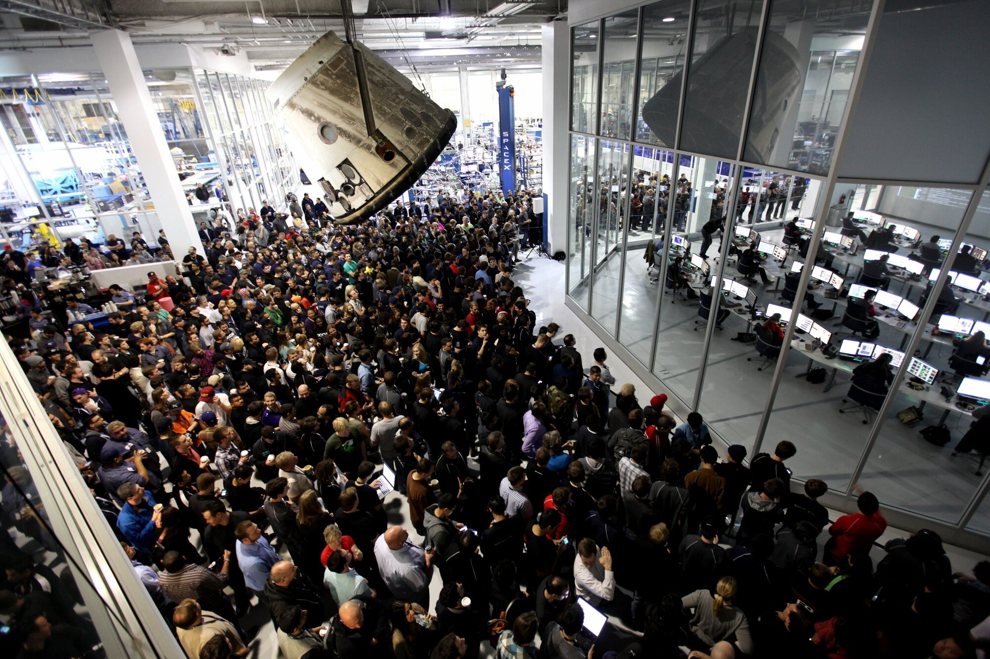 A large crowd of spacex employees inside the hawthorne california headquarters with a spacecraft module hanging from the ceiling and glass enclosed control rooms visible in the background