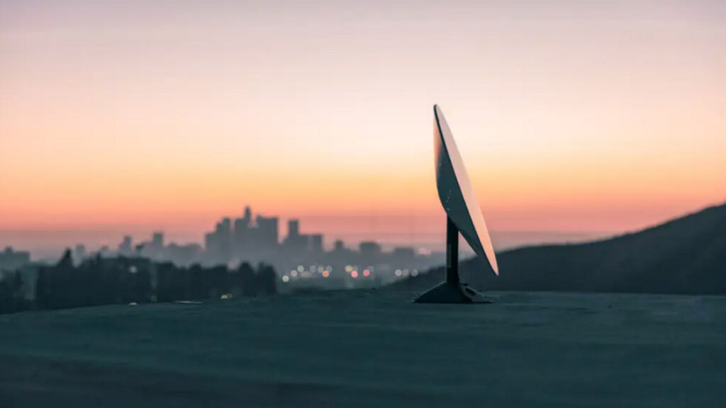 A starlink satellite dish sits on a hilltop overlooking a city skyline at sunset with the sky painted in warm orange and pink hues