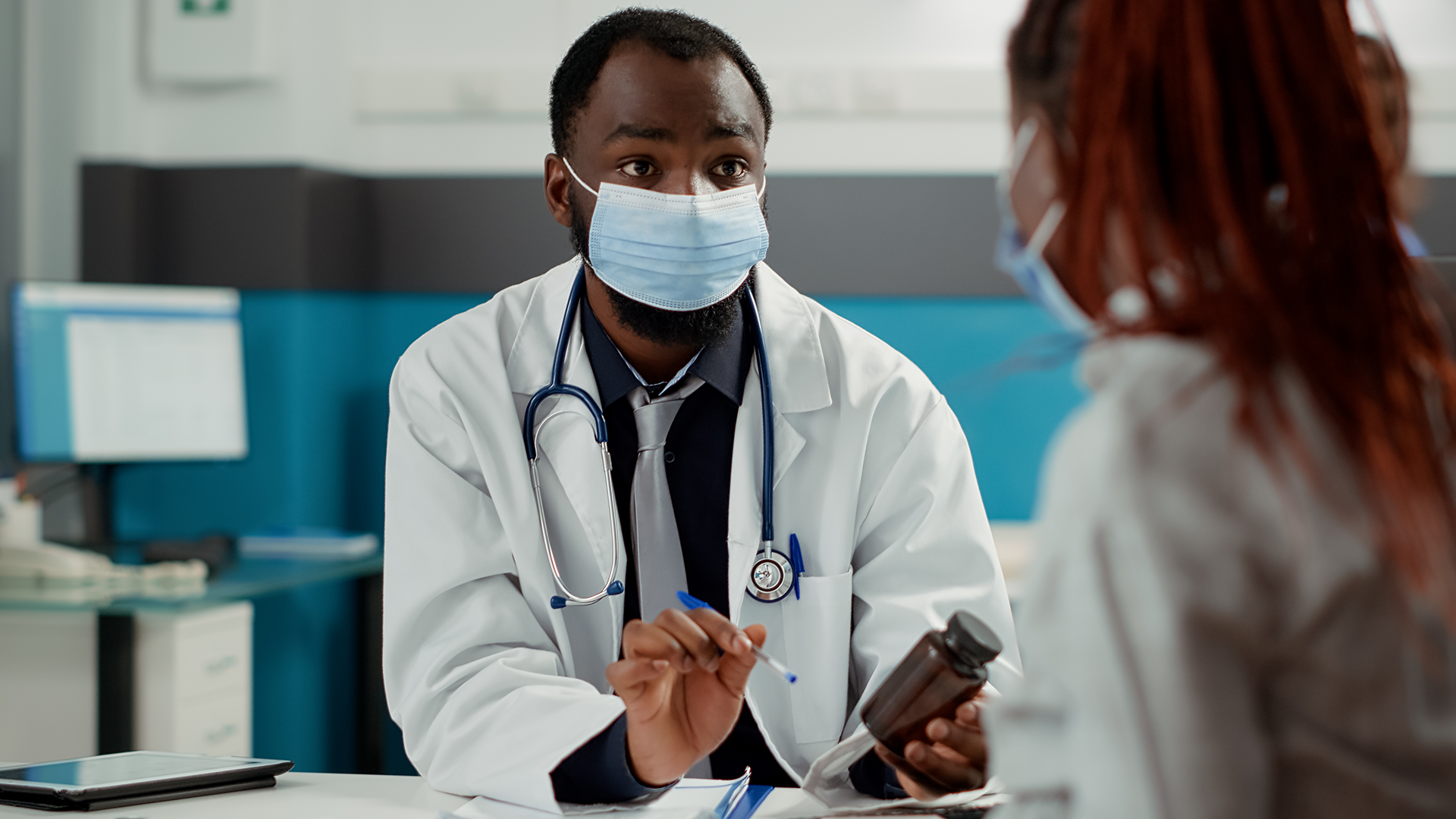 Male doctor wearing a white coat and face mask speaks to a masked female patient while holding a medication bottle in a health clinic
