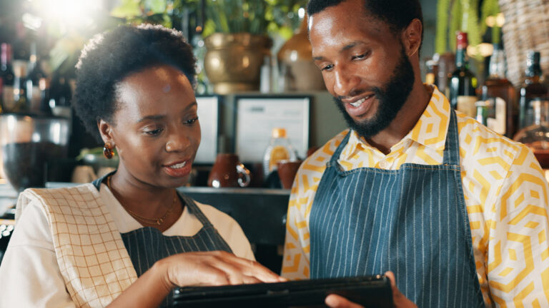 African man and woman wearing aprons looking at a tablet while working together in a restaurant setting.