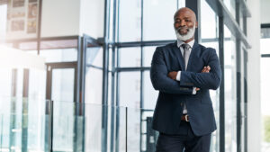 A mature businessman with a gray beard dressed in a navy suit and tie standing with crossed arms in a modern glass office