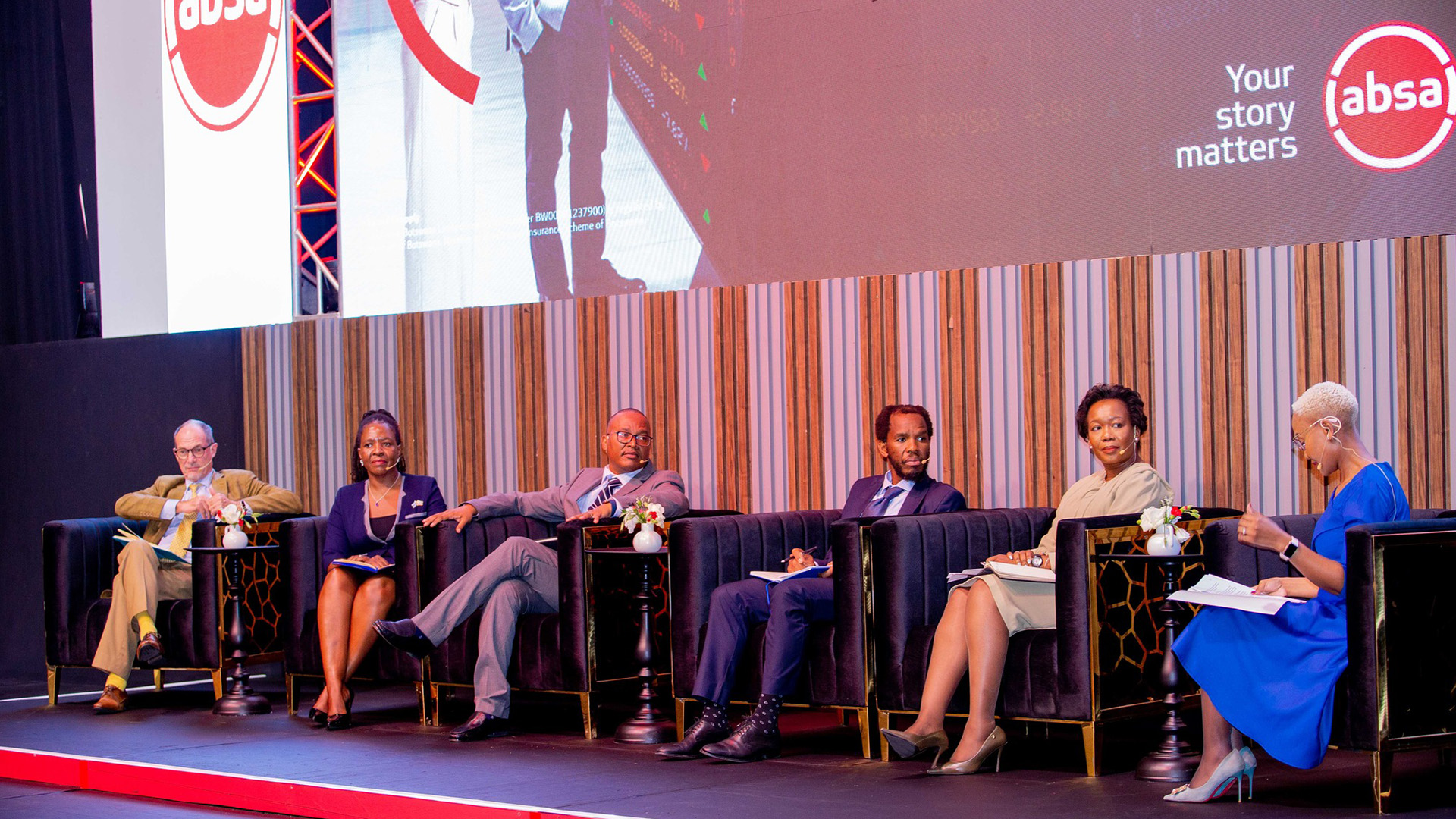 Absa-Your Story Matters-Executive Panel | Brimco Six panelists seated in dark armchairs on a stage in front of a large screen displaying the absa logo and the slogan your story matters