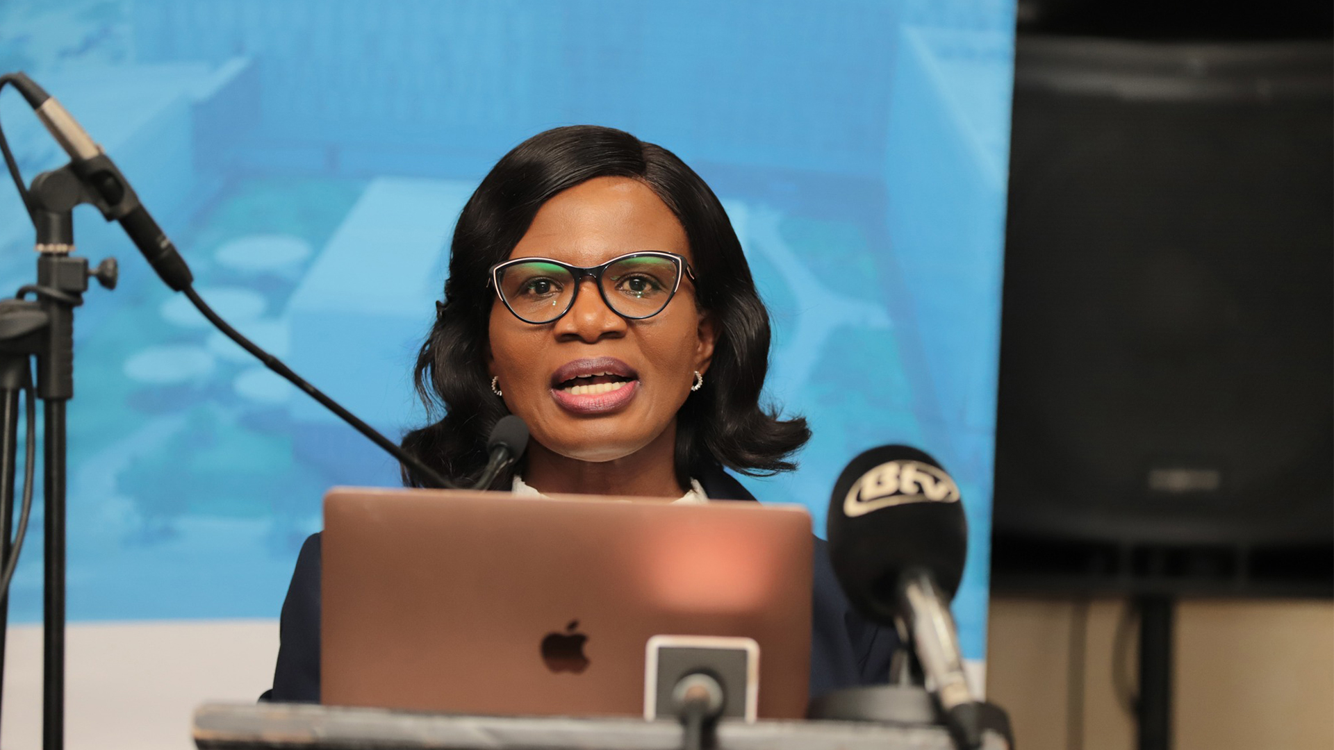 BURS Commissioner General Jeanette Makgolo Addresses Revenue Forum | Brimco Jeanette makgolo wearing glasses and a dark blazer speaks into a microphone behind a podium with an open laptop and additional microphones a blue presentation screen behind her