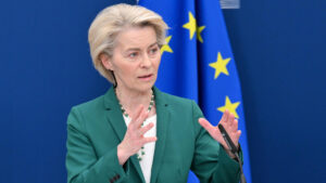 Ursula von der leyen gesturing while speaking in front of an eu flag backdrop during a press briefing