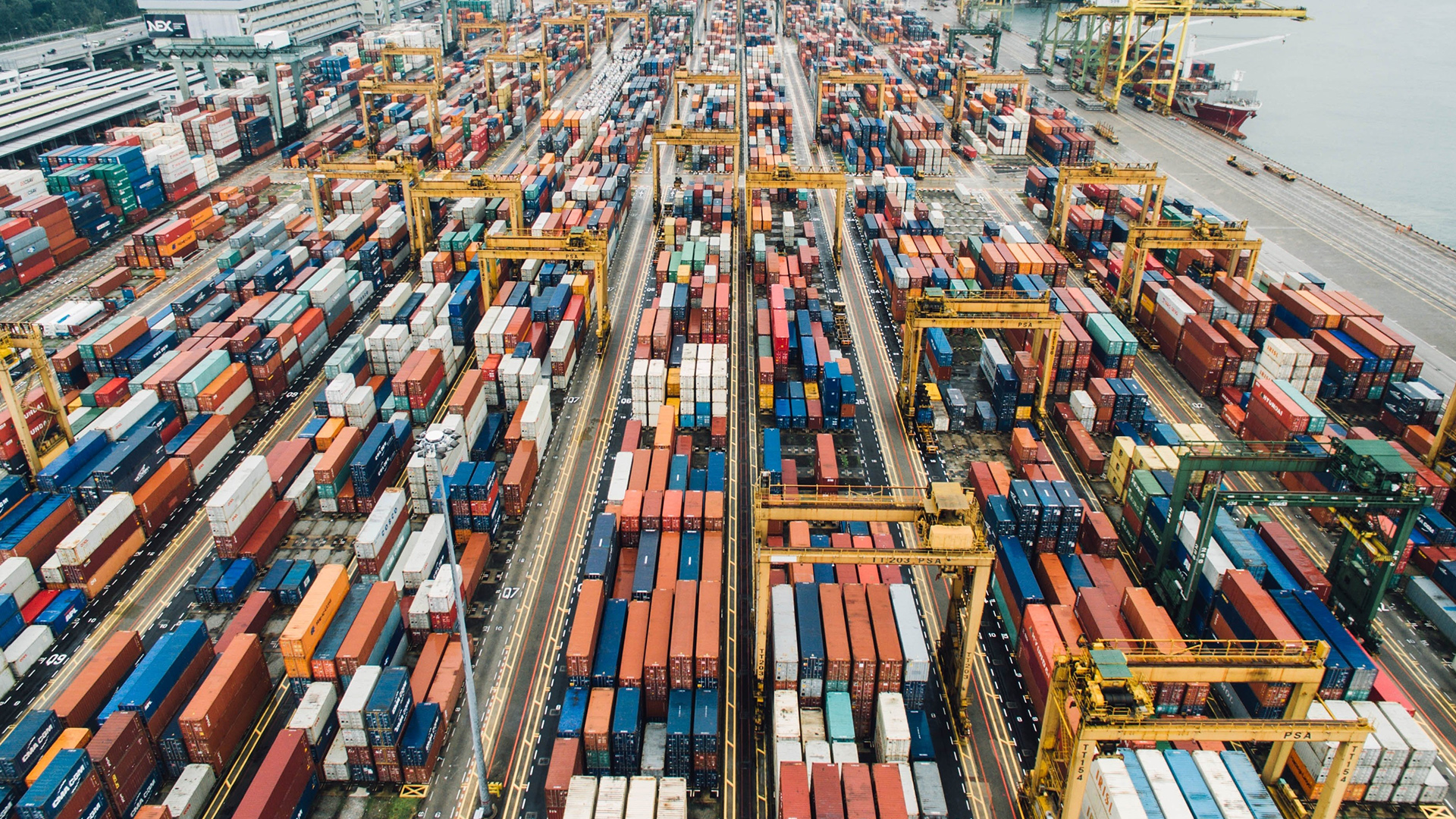 Global Trade in Motion Aerial View of a Busy Container Port | Brimco Aerial photograph of a container port showing hundreds of stacked shipping containers in neat rows multiple yellow gantry cranes and a docked cargo ship