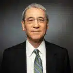 Gordon chang in a dark suit and striped tie facing the camera with a neutral expression against a dark background