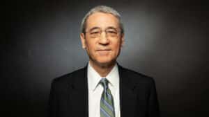 Gordon chang in a dark suit and striped tie facing the camera with a neutral expression against a dark background