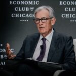 Jerome Powell speaking at the Economic Club of Chicago, seated with a binder in hand and a microphone on his suit.