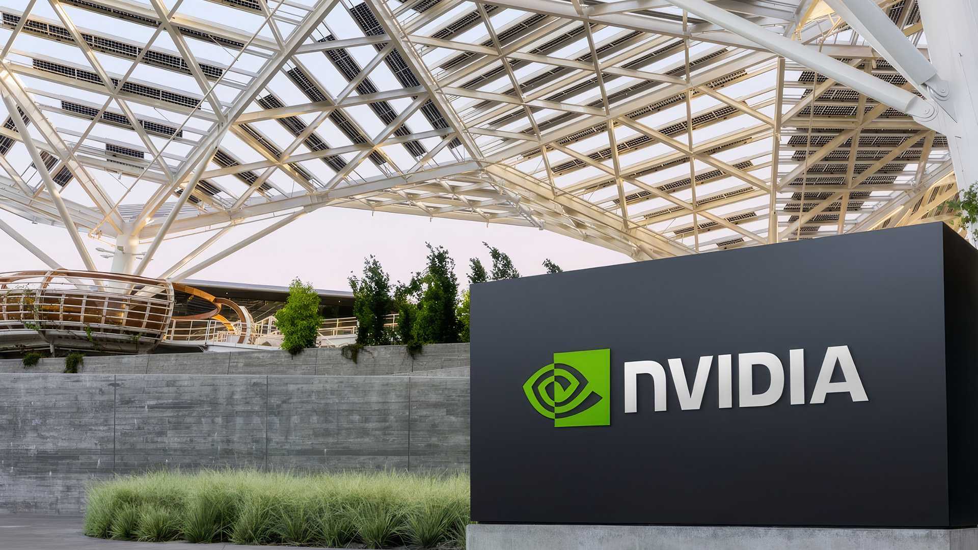 NVIDIA Headquarters Under Solar Canopy | Brimco A black sign with the green nvidia logo stands before a concrete wall and grasses with a geometric metal and solarpanel canopy overhead at nvidias campus entrance