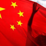 Shadowed Hands Behind the Chinese Flag | Brimco Silhouette of hands reaching up behind a translucent chinese flag fluttering against a partly cloudy sky