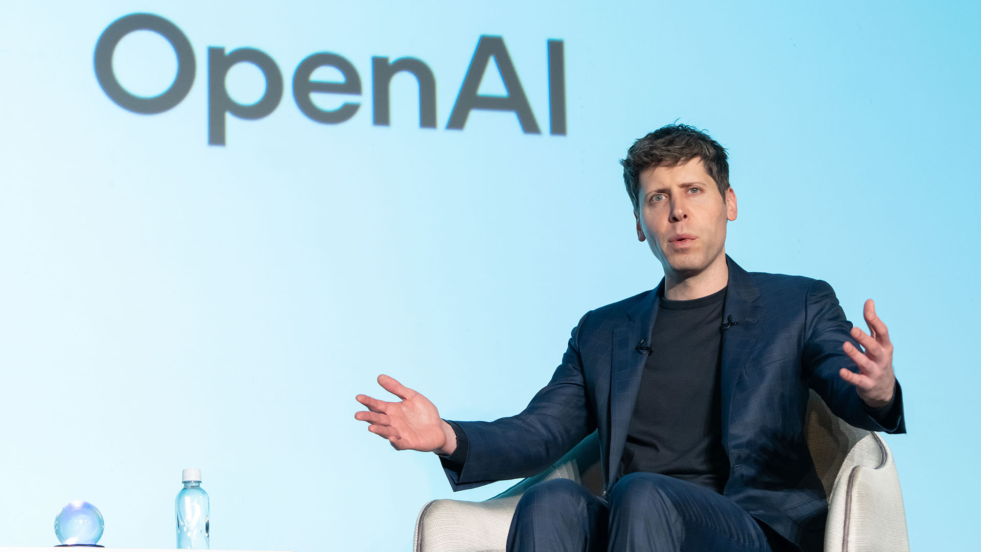 Shaping the Future A Conversation on AI at OpenAI | Brimco A man in a navy suit speaking passionately on stage with an openai logo in the background