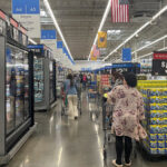 Shoppers with carts walk down a brightly lit supermarket aisle past freezer cases and pallet stacks of drinks with large rollback price signs overhead and aisle markers for desserts frozen pizza and snacks