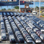 A lone port worker in blue overalls stands among dozens of neatly lined rows of new cars parked on an export dock with a container ship and cranes in the background