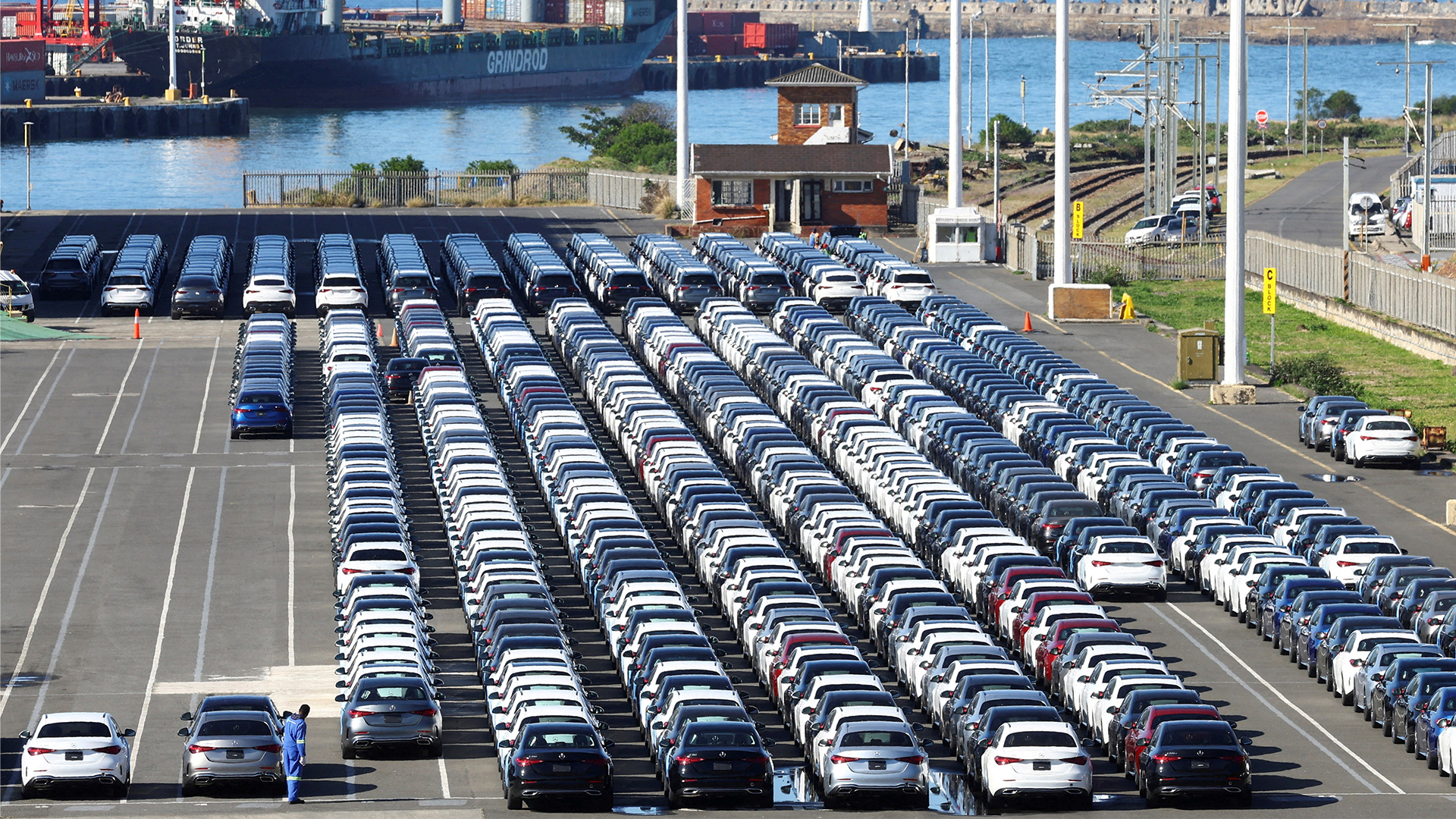 South African Port Stockpiles New Cars for Export | Brimco A lone port worker in blue overalls stands among dozens of neatly lined rows of new cars parked on an export dock with a container ship and cranes in the background