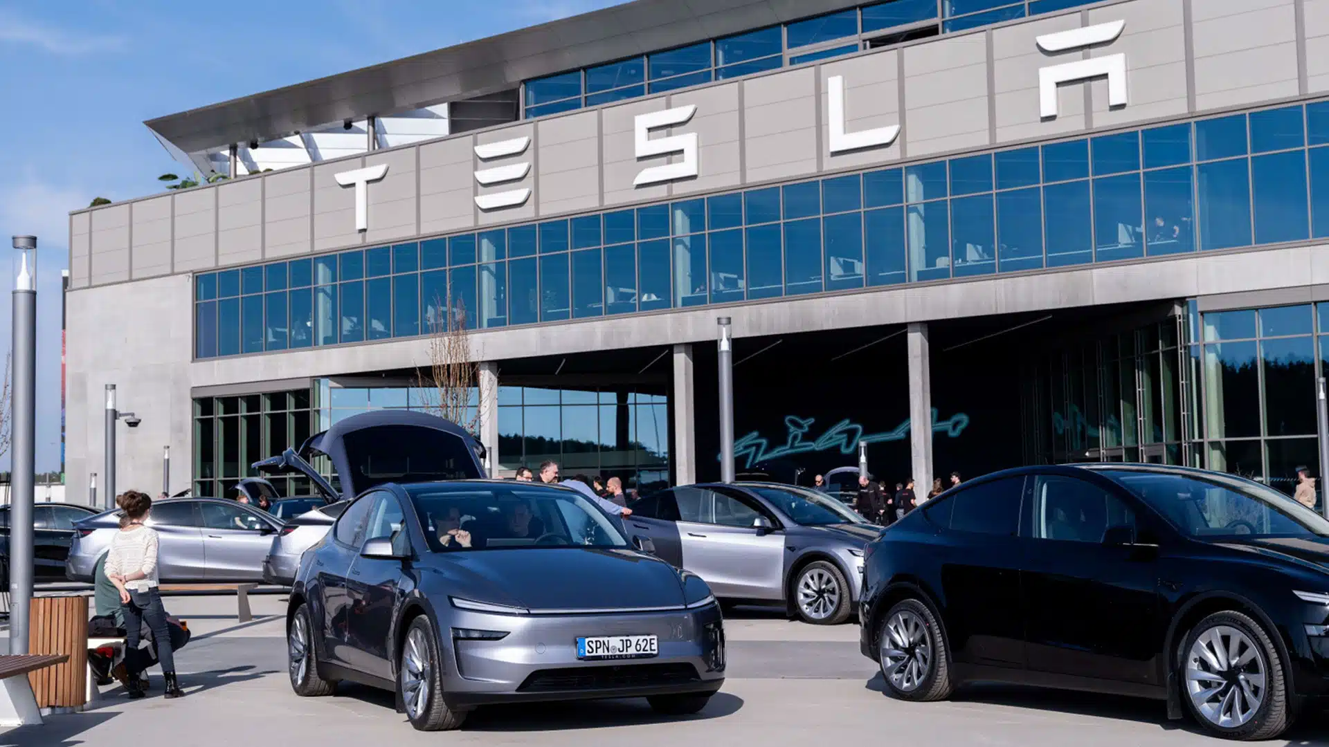 Tesla electric vehicles parked outside a tesla gigafactory with people walking and interacting near the cars