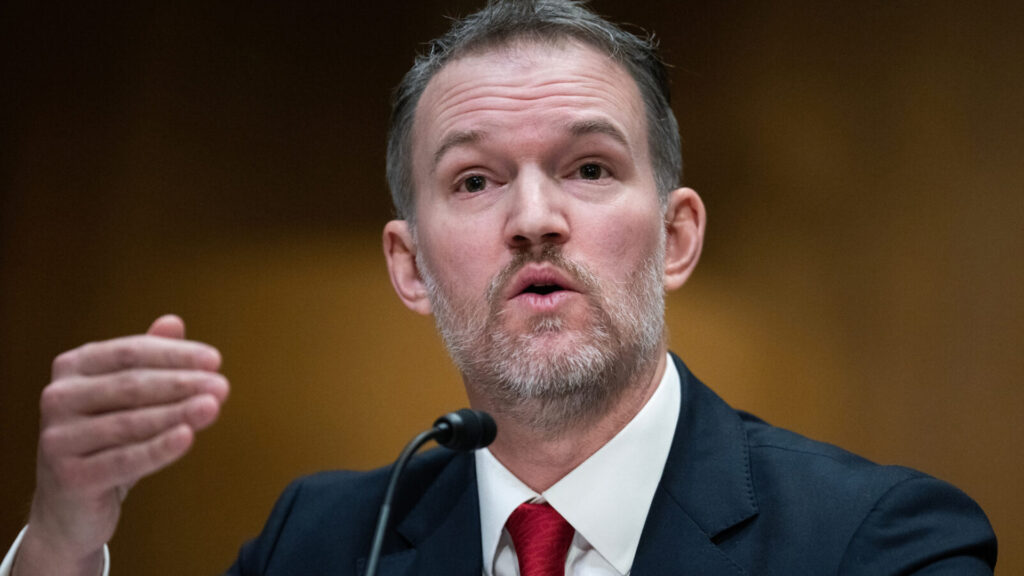 Greer speaking into a microphone at a senate hearing wearing a dark suit and red tie gesturing with his hand