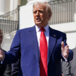 Donald trump in a dark blue suit and red tie stands on the white house lawn hands outstretched as he speaks with two suited aides standing behind him
