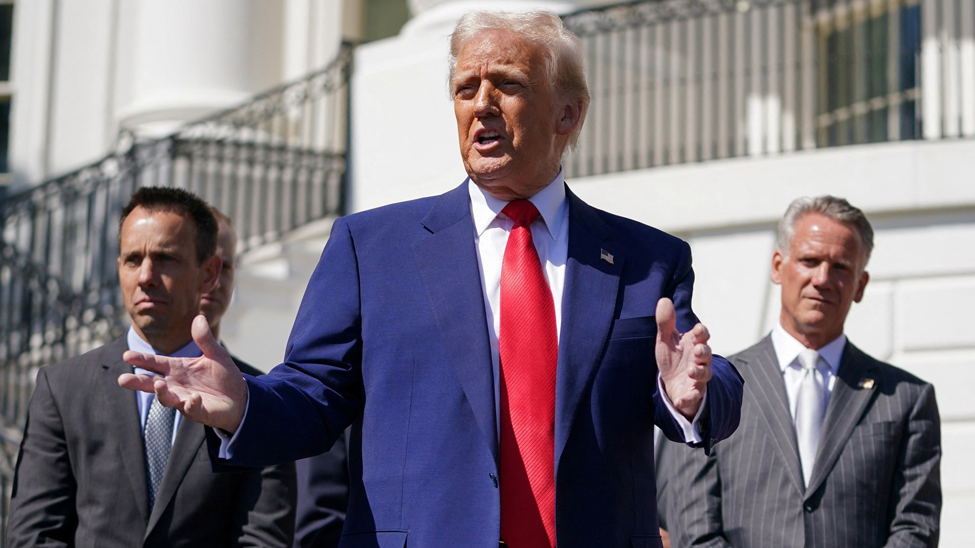 Trump Holds Press Briefing on White House Lawn | Brimco Donald trump in a dark blue suit and red tie stands on the white house lawn hands outstretched as he speaks with two suited aides standing behind him