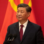 Xi jinping in a dark suit and red tie stands at a lectern with dual microphones with a large red flag bearing the yellow hammer and sickle behind him