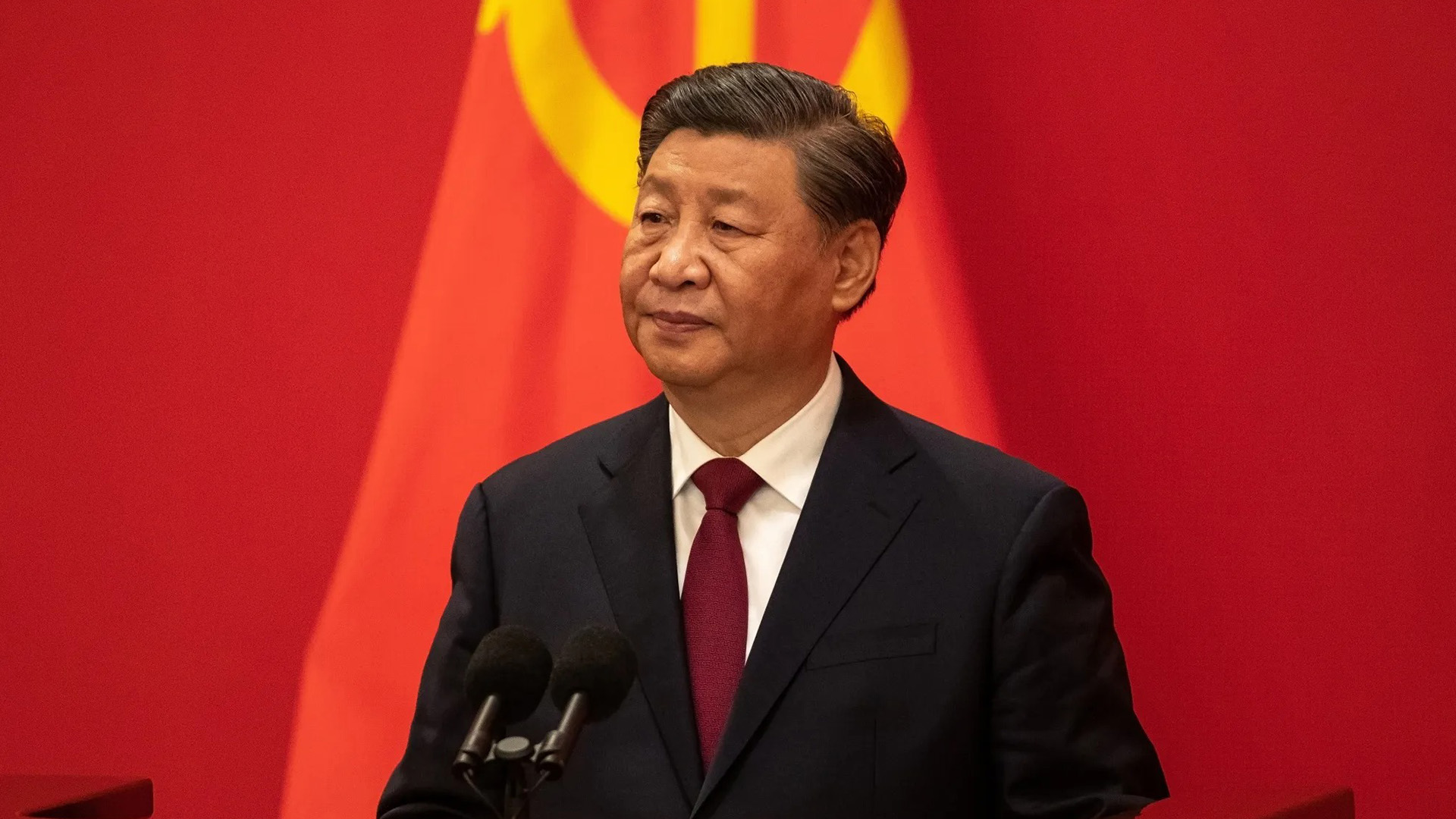Xi Jinping Delivers Address at Party Congress | Brimco Xi jinping in a dark suit and red tie stands at a lectern with dual microphones with a large red flag bearing the yellow hammer and sickle behind him