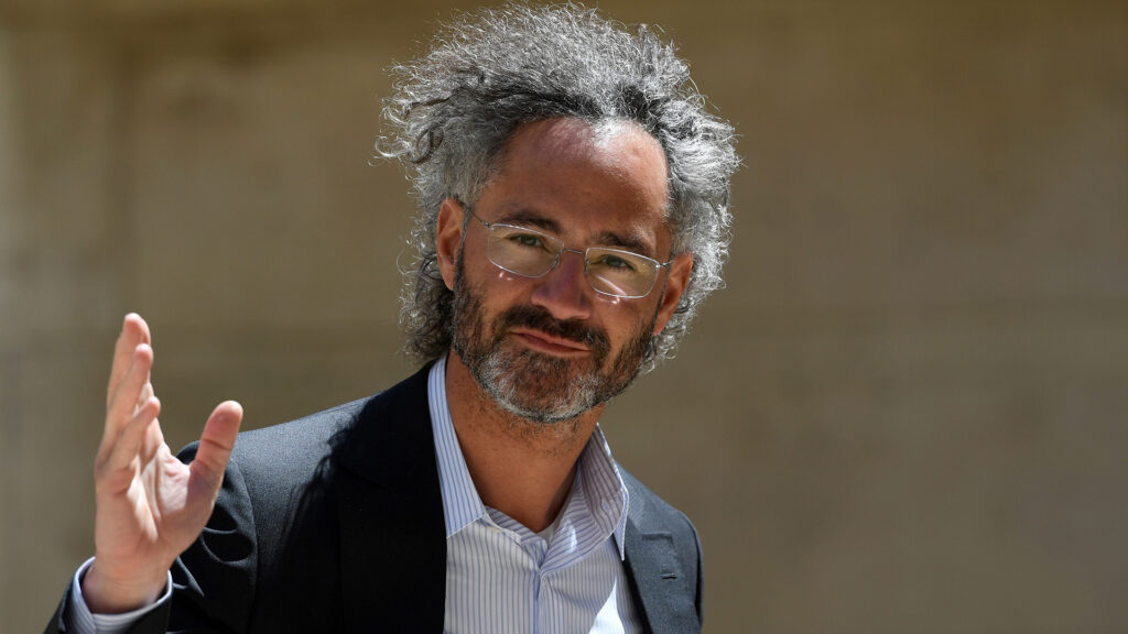 Alex karp ceo of palantir technologies wearing glasses and a suit raising his hand in a thoughtful gesture