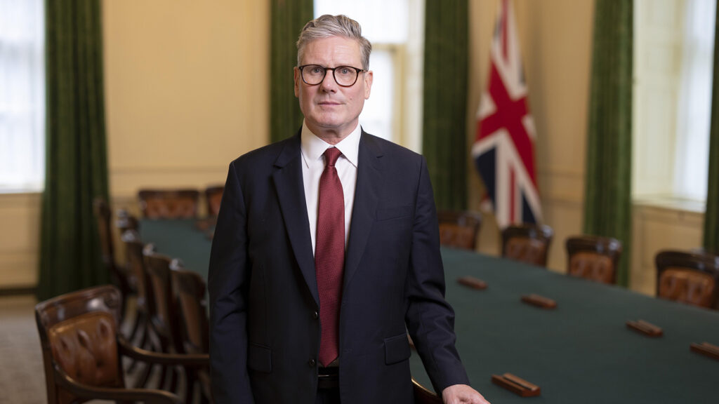 Keir starmer wearing a dark suit and red tie standing in a formal meeting room with a long table and the union jack flag behind him