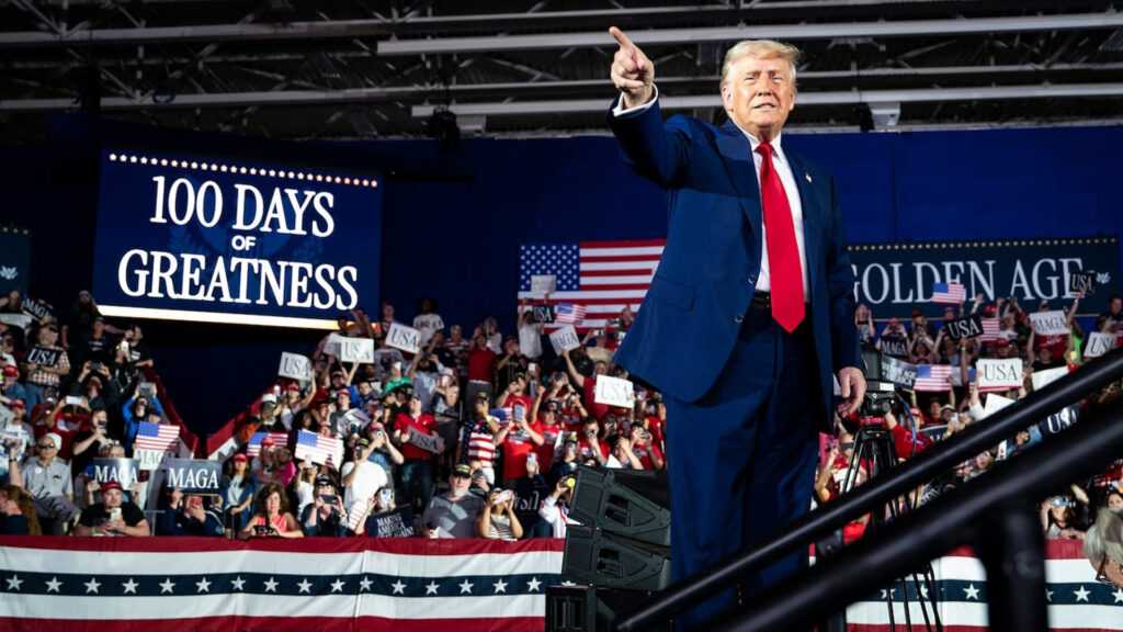 Donald trump points toward the crowd at a 100 days of greatness rally with supporters waving american flags and signs reading usa and maga 