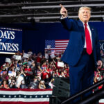 Donald Trump points toward the crowd at a "100 Days of Greatness" rally, with supporters waving American flags and signs reading “USA” and “MAGA.”