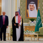 Donald Trump and Crown Prince Mohammed bin Salman standing side by side at the Saudi Royal Palace, with the Saudi flag and a portrait of King Salman in the background.