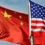 Close-up image of the United States and China flags waving on flagpoles against a blue sky.