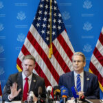 U.S. Trade Representative Jamieson Greer and Secretary of the Treasury Scott Bessent speaking at a press conference in Geneva with U.S. flags and microphones in the foreground.