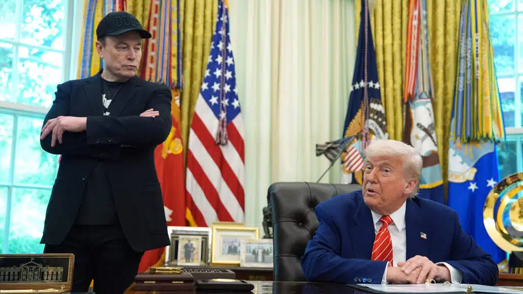 Elon musk stands with arms crossed while president donald trump sits at the resolute desk in the oval office during a discussion