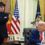 Elon musk stands with arms crossed while president donald trump sits at the resolute desk in the oval office during a discussion