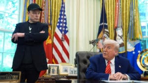 Elon musk stands with arms crossed while president donald trump sits at the resolute desk in the oval office during a discussion