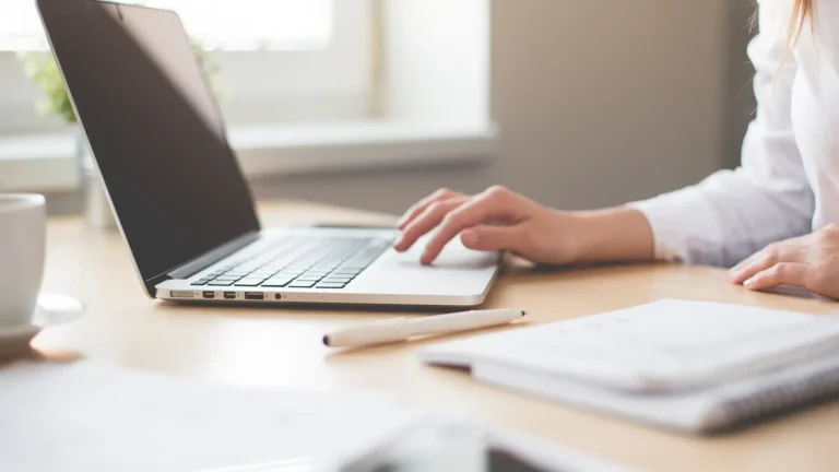 Close up of a womans hands using a laptop at a wooden desk with documents and a coffee cup nearby