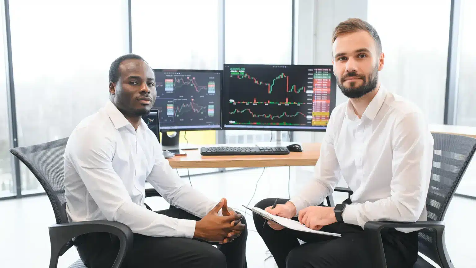 Two professional brokers in white shirts seated at a trading desk with multiple monitors displaying real time stock charts and financial data