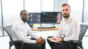 Two professional brokers in white shirts seated at a trading desk with multiple monitors displaying real time stock charts and financial data