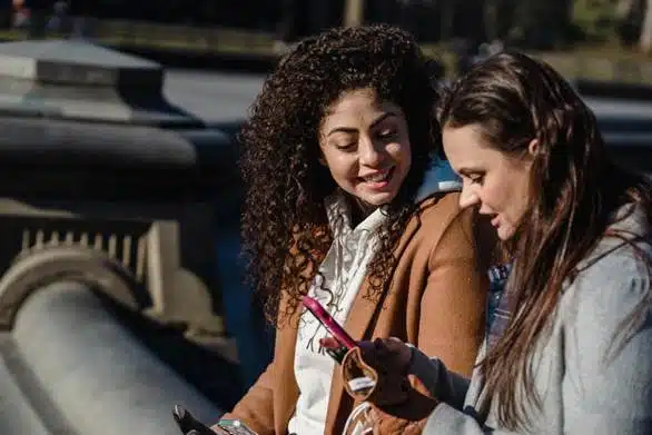 Two smiling women looking at a pink phone outdoors while sitting on a stone structure