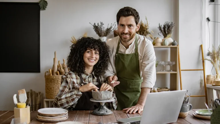 Smiling pottery studio owners working on ceramic art near a laptop