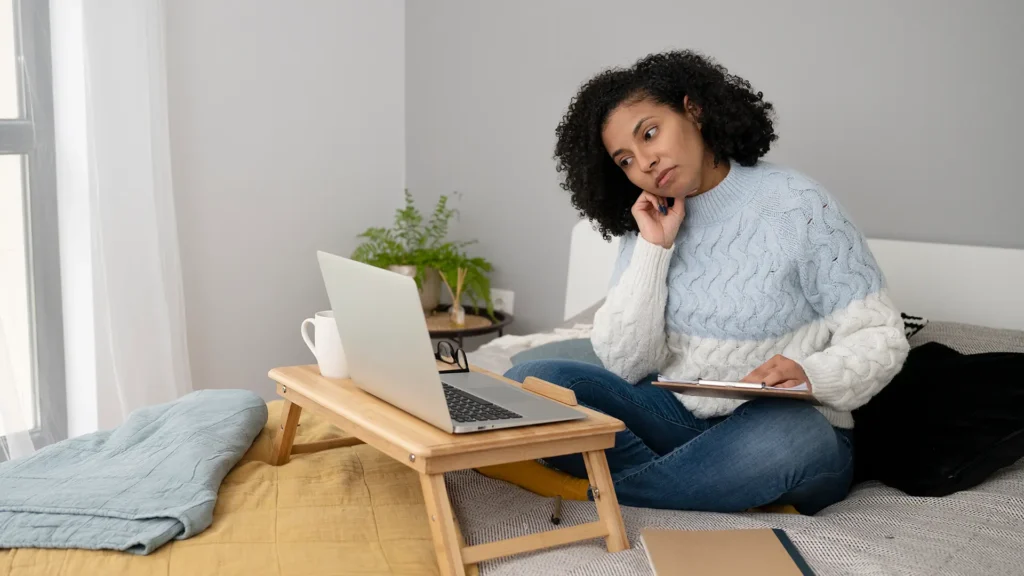 Woman in casual clothing sitting on a bed and attending a remote training session via laptop while taking notes