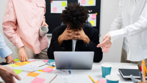 Stressed employee covering face while colleagues discuss around laptop