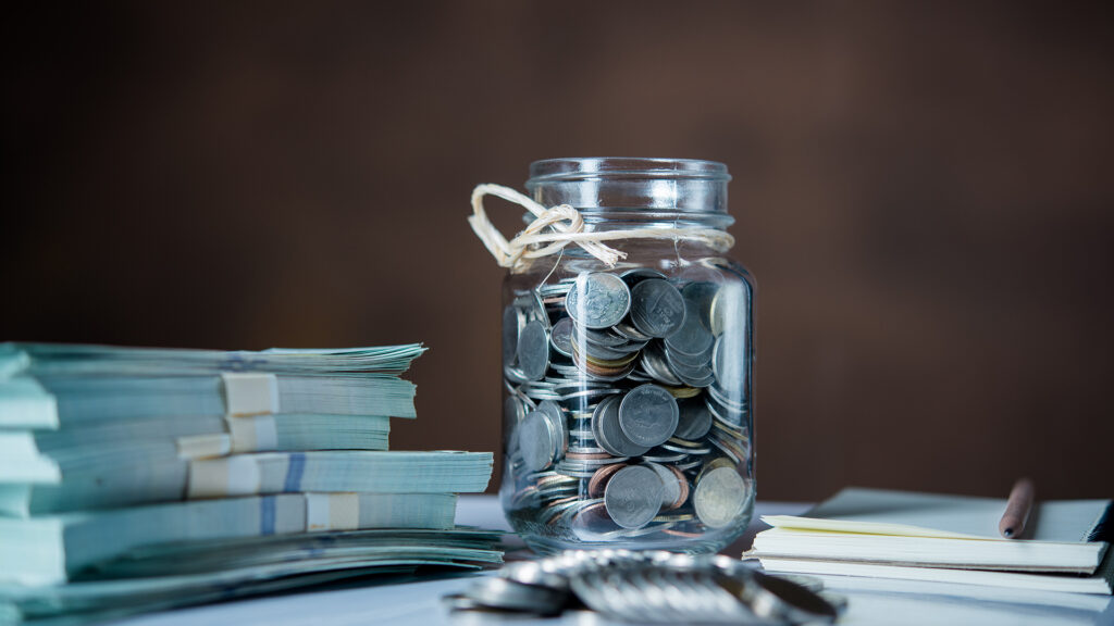 Glass jar filled with coins placed beside bundles of cash, a notebook, and a pen, representing saving and financial planning.