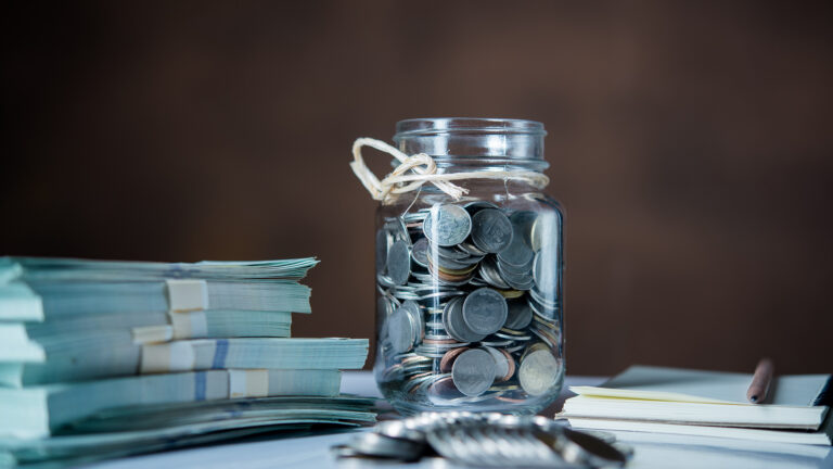 Glass jar filled with coins placed beside bundles of cash a notebook and a pen representing saving and financial planning