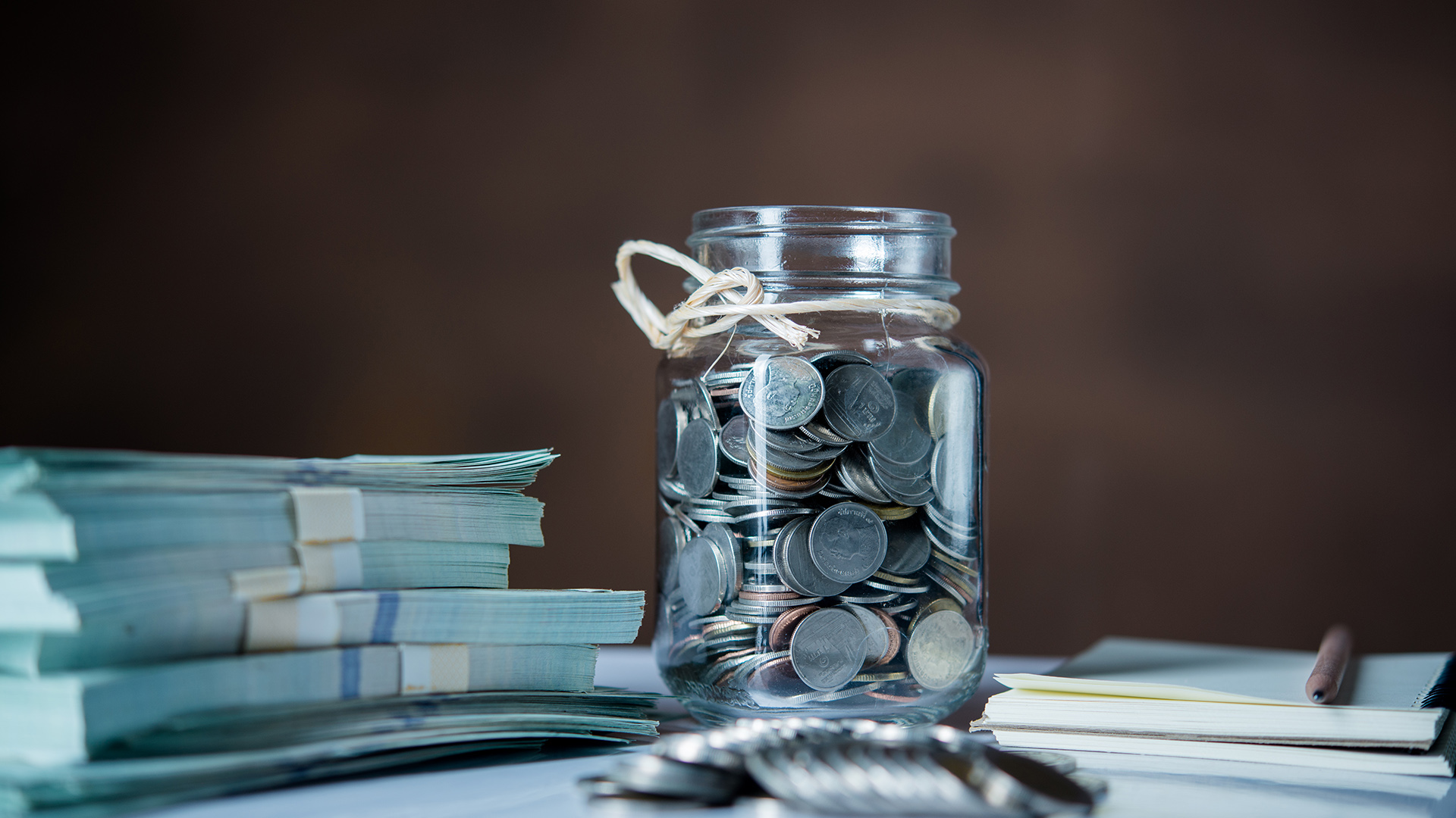 Glass jar filled with coins placed beside bundles of cash a notebook and a pen representing saving and financial planning