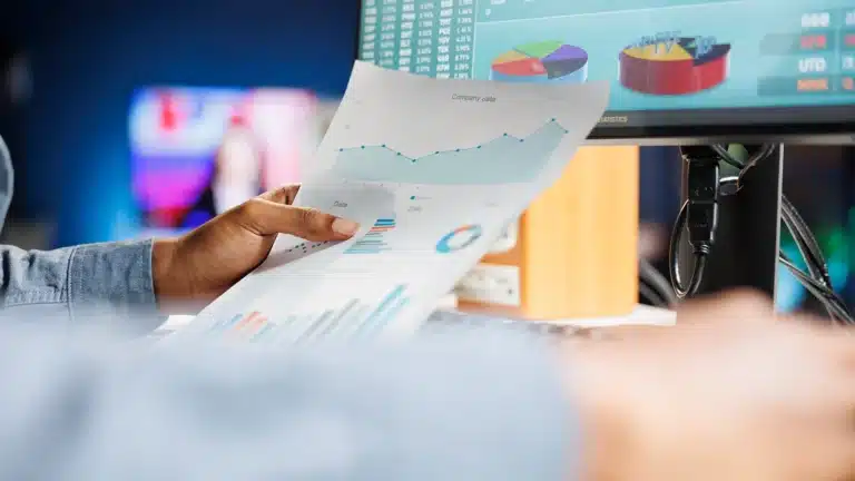 Business consultant analyzing financial charts on paper while reviewing data visualizations on a computer screen.