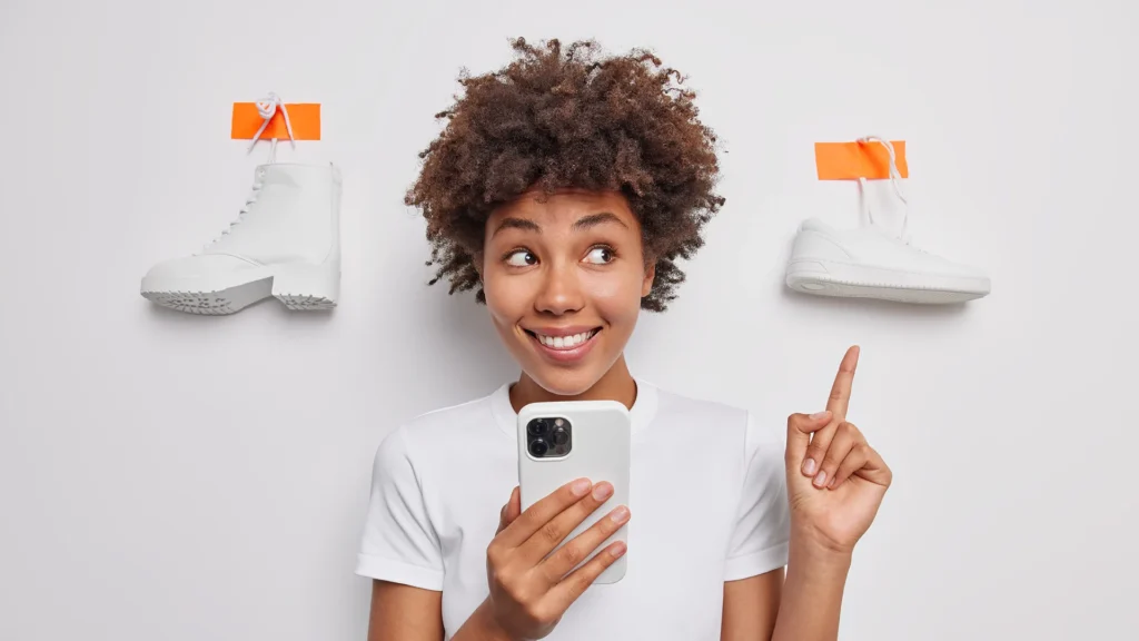 Smiling woman holding a smartphone choosing between two pairs of shoes online.