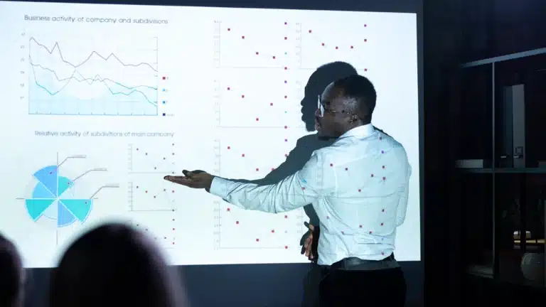 African businessman presenting company performance graphs and data visualizations on a projector screen during a meeting.
