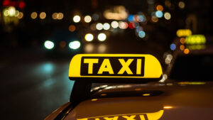 Close up of a yellow illuminated taxi sign on a car roof at night with blurred city lights in the background
